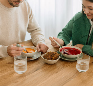 Cuplu savurând supe cremă sănătoase, una de sfeclă roșie și alta de morcov, alături de crutoane crocante și apă, la o masă din lemn.